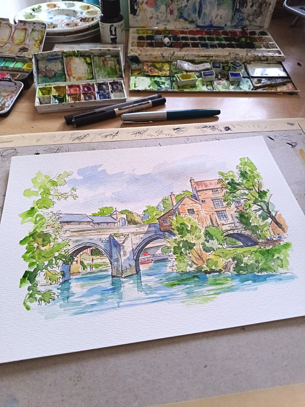 A watercolor painting of a river scene with stone arches, green trees, and buildings, displayed on a table surrounded by paint palettes, brushes, and markers.