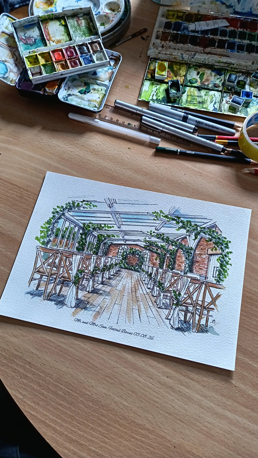 Watercolor illustration of a wedding venue interior with wooden chairs and greenery on a wooden desk with art supplies