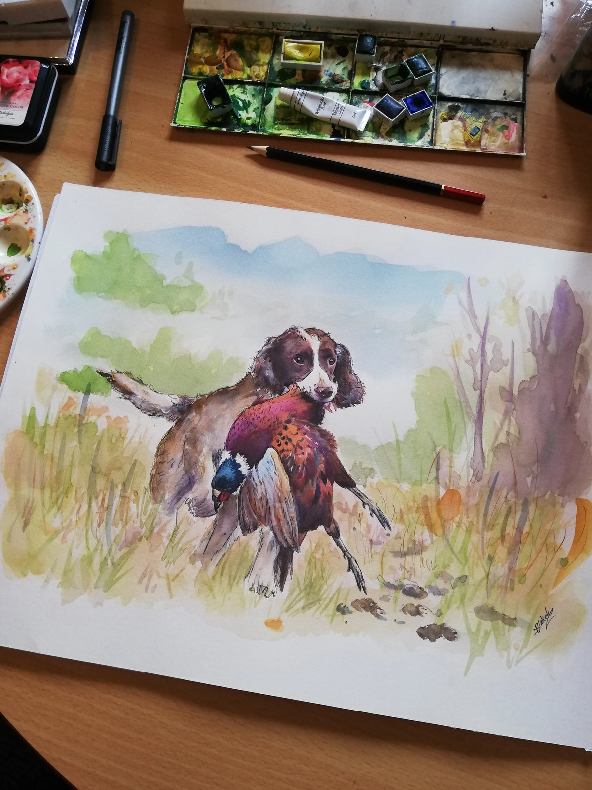 A watercolor painting of a hunting dog carrying a pheasant in its mouth, displayed on a wooden table with an artist's watercolor palette, paint tubes, and brushes.