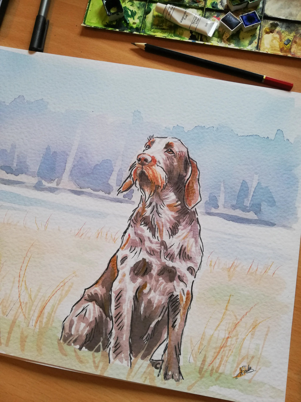 A watercolor painting of a brown and white spotted dog sitting in a grassy field with a blue water background, shown on a wooden table with paint tubes, a paintbrush, and a marker nearby.