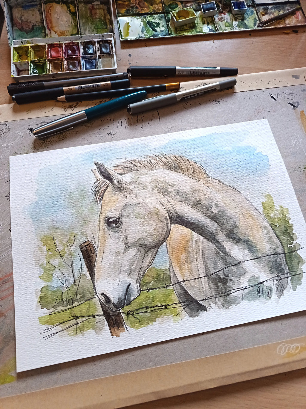 A watercolor painting of a light gray horse on textured paper, displayed on a wooden table alongside watercolor palettes, paintbrushes, and markers.