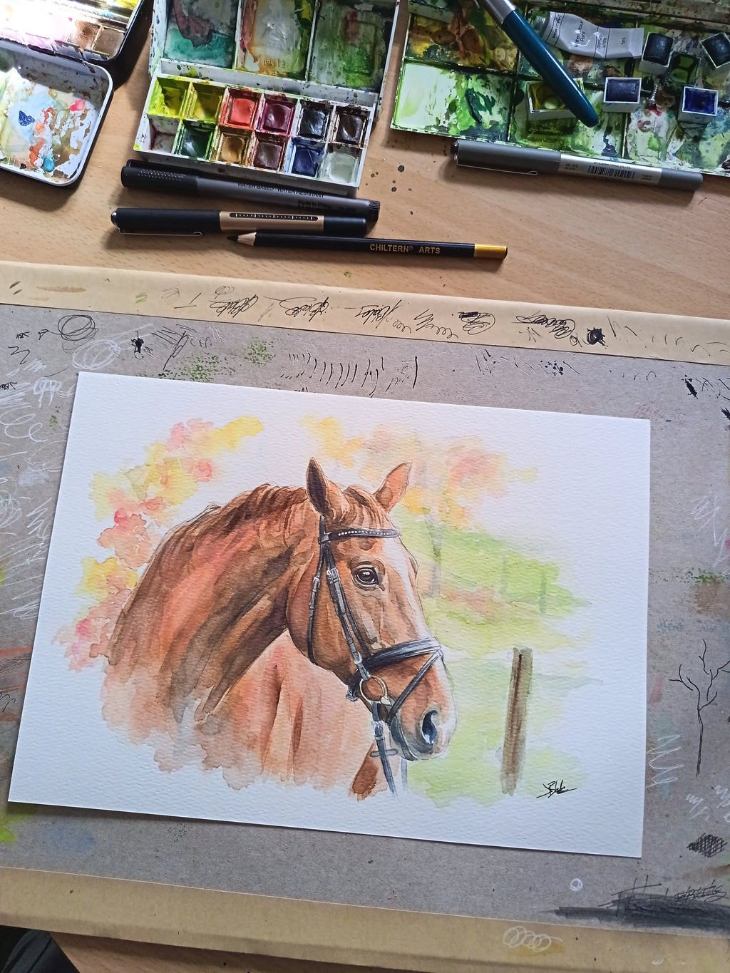 A watercolor painting of a brown horse with a bridle, shown on a sheet of white paper laid on a wooden artist's table surrounded by paint palettes, pencils, and brushes.
